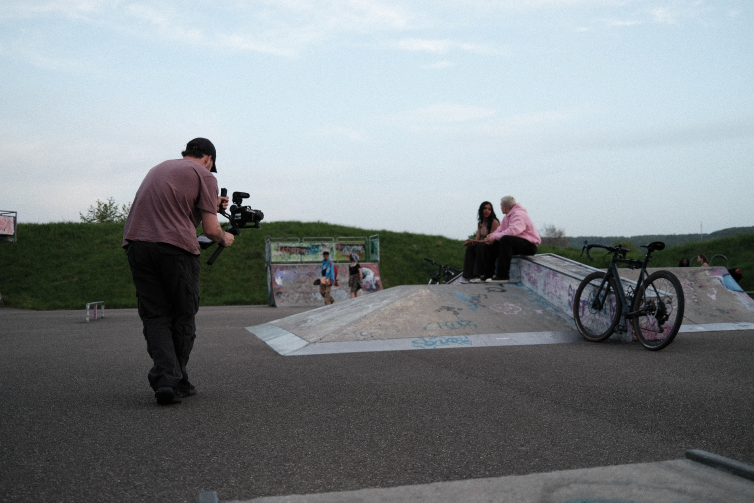 Making of scene showing a camera man with a gimbal walking thowards two young actors sitting on a skate ramp